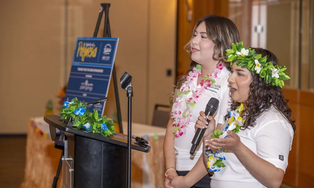 Ale & Evy wearing white and flowers speaking at an event in a mic