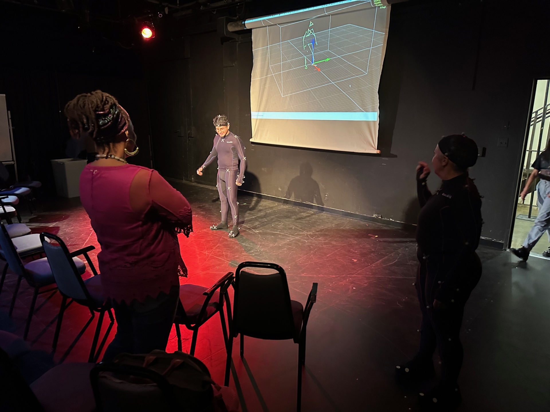 Student in a motion capture suit in a black wall studio with two other instructors watching. There is a screen behind the student showing the avatar animation.