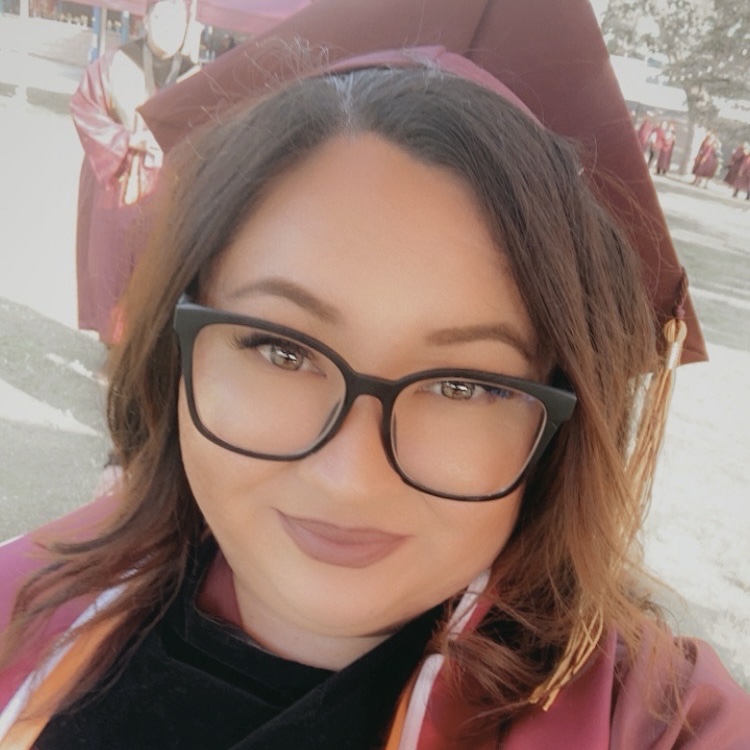 Mickela is smiling at the camera wearing a graduation cap and gown. Mickela has light red colored hair and light brown eyes.