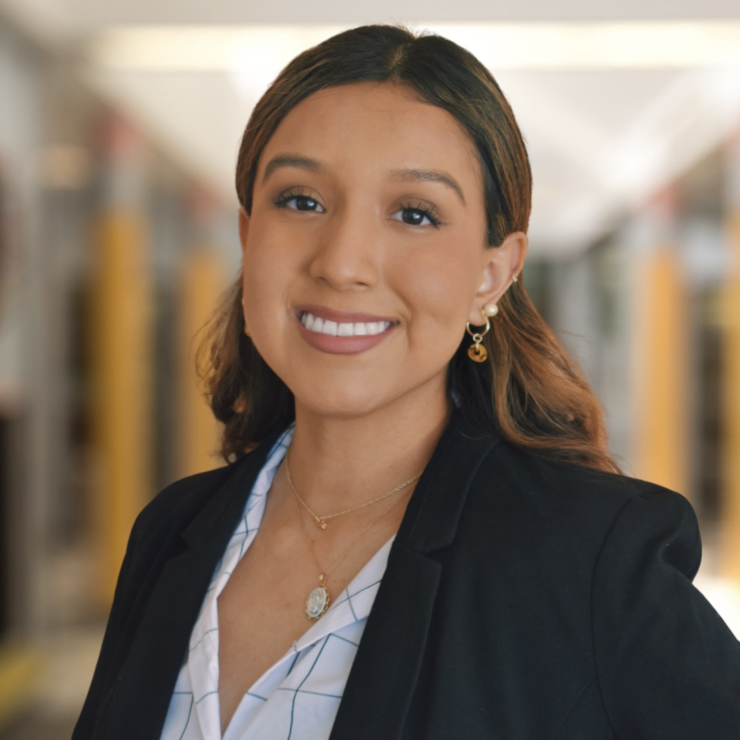 The image shows a picture of Ariana Cardoso. She has brown hair and dark eyes, she is outside smiling at the camera. She is wearing a black jacket and grid button up top.