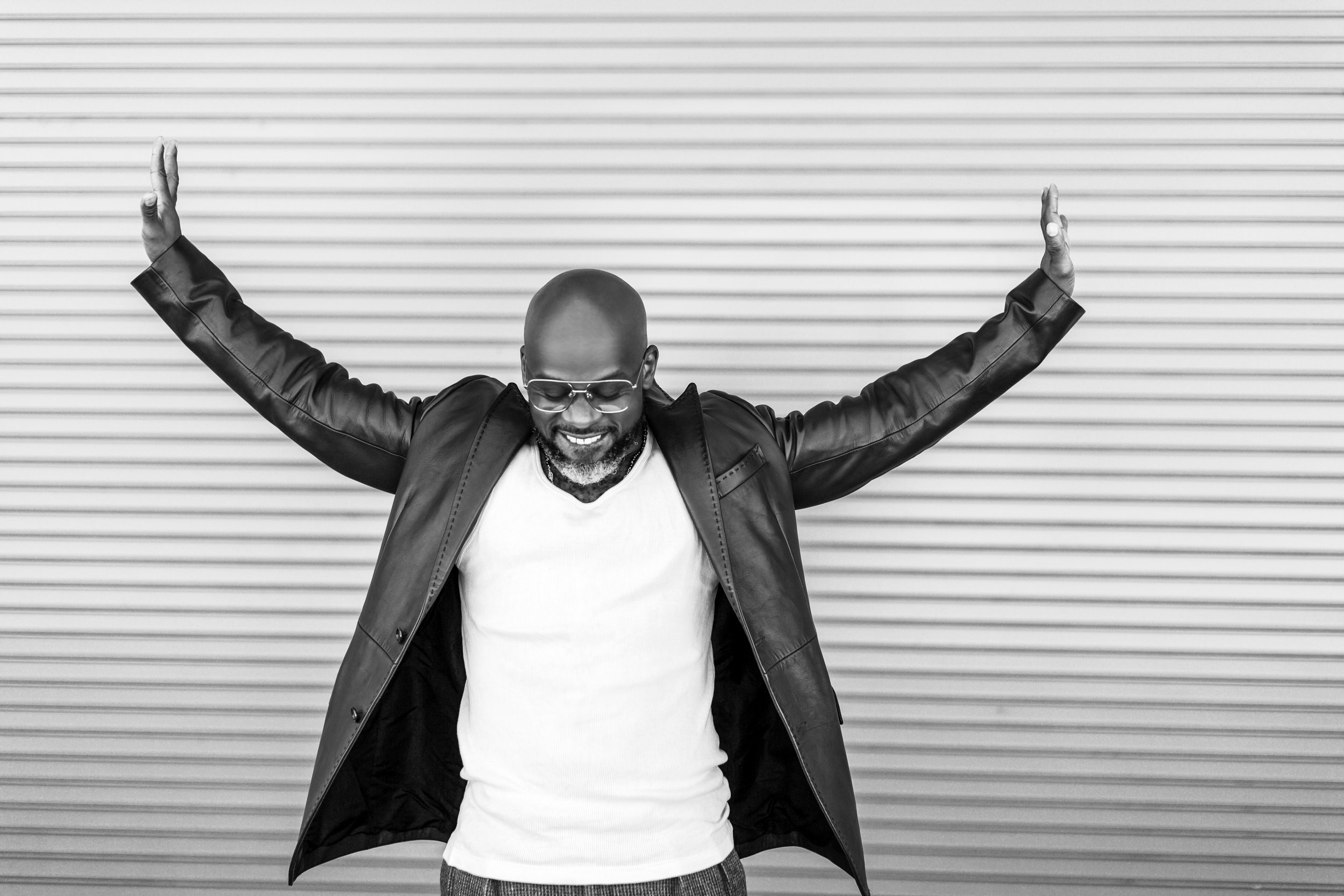 Marc Bamuthi Joseph, black and white image, African American man, shaved head, looking down and smiling, arms outstretched, leather jacket and white shirt