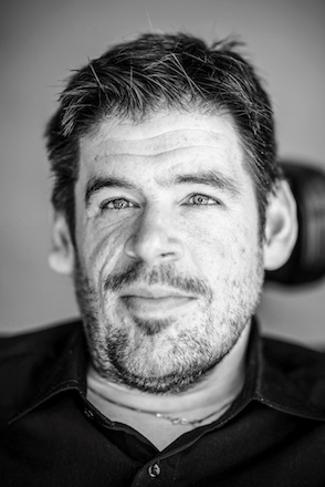 Black and white image of white man looking at camera, stubly beard, short dark hair, dark collared shirt and gold chain