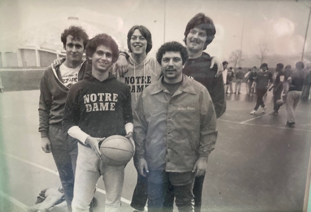Five young men, some in Notre Dame shirts, stand smiling in front of a basketball court before the game. Pedro Santoni holds the ball in front.