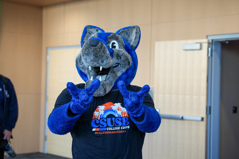 Cody the Coyote wearing a CSUSB College Corps t-shirt and making the "yotes up" gesture