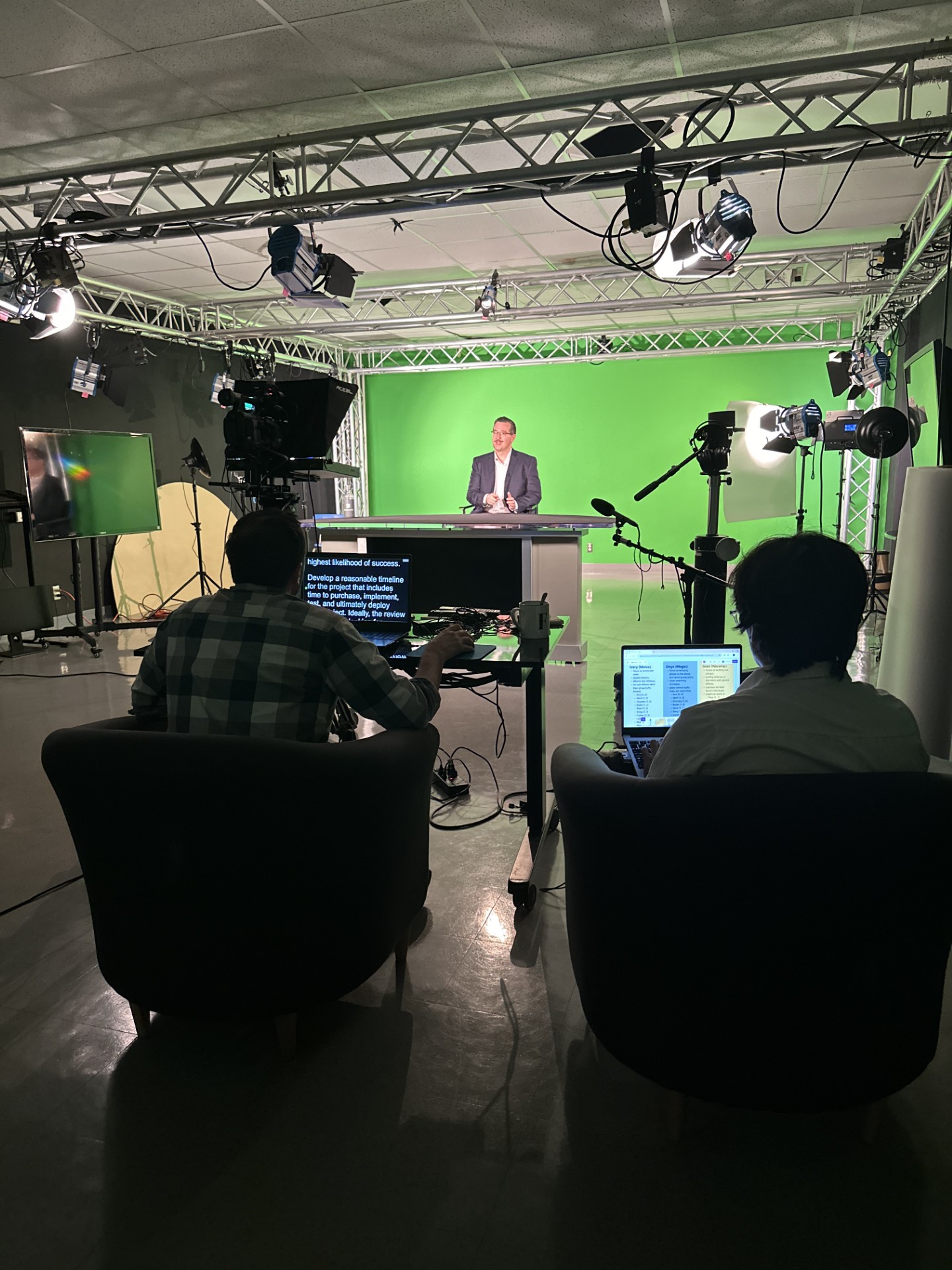 A live video production in progress inside a green screen studio. A man in a blazer is seated at a desk in front of a bright green chroma key backdrop, lit by multiple overhead and side studio lights. In the foreground, two operators sit in armchairs; one controls a teleprompter displaying text, while the other works on a laptop showing what appears to be a script or production notes. A camera with a teleprompter attachment is positioned between the crew and the presenter. The studio setup includes lighting trusses, monitors, microphones, and cabling arranged for a professional shoot.