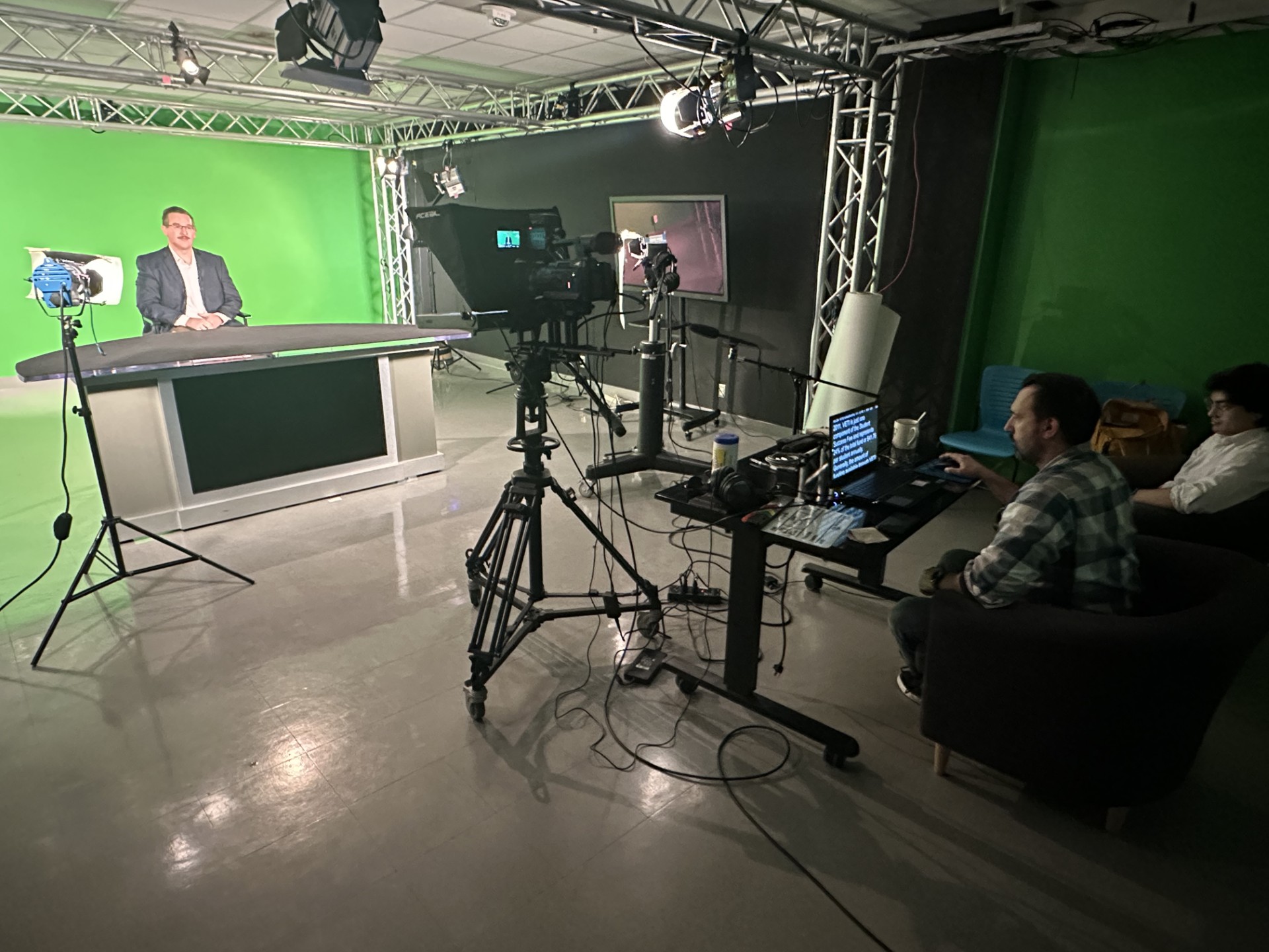 A behind-the-scenes view of a green screen video production studio. A man in a suit is seated at a modern news-style anchor desk in front of a large green screen backdrop. Multiple professional studio lights and cameras are set up around him, including a main camera with a teleprompter. In the foreground, a technician operates a laptop displaying the teleprompter script while seated next to another person observing. Cables, microphones, and production equipment are organized on rolling carts. The room is framed with a metal truss lighting rig and features studio walls painted partially black and green.