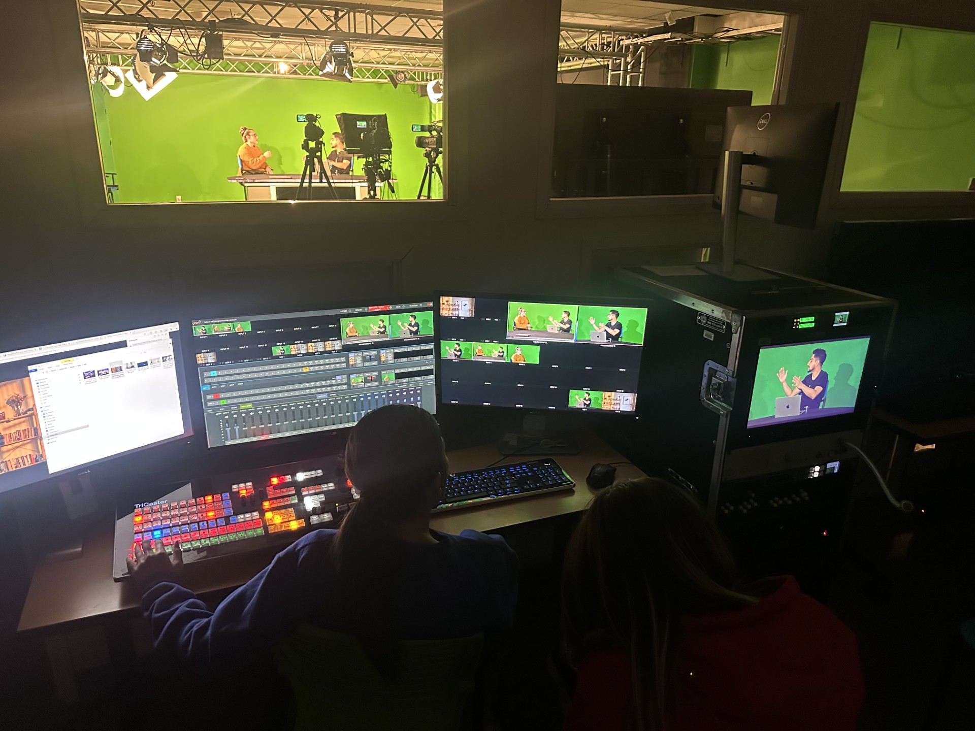 A production in progress inside the PL-056 studio at California State University, San Bernardino. The image shows a control room with two operators managing a live video shoot. Multiple monitors display a green screen set with two presenters standing behind a news-style desk, visible through a window separating the studio and control room. The control desk features a professional video switcher, audio mixer, and editing software. Various live camera feeds appear on the screens, including chroma-keyed previews. One rack-mounted monitor shows a close-up of a presenter with graphics keyed in. The studio setup includes studio lights on a truss system and multiple cameras on tripods.
