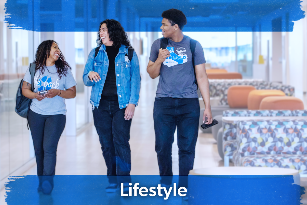 Three young people walk and chat in a bright hallway, smiling and relaxed. They wear casual clothes and backpacks. The mood is friendly and cheerful.