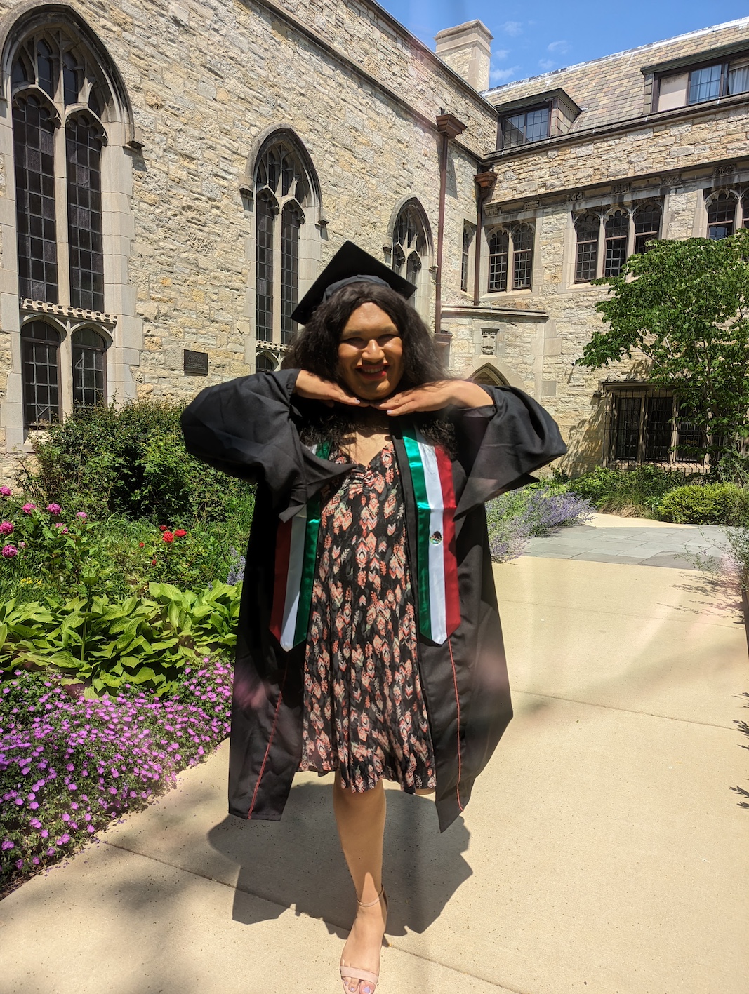 Nayeli Gonzalez, in graduation regalia with Mexican flag sash, standing in front of stone building hands under chin and smiling widely