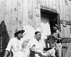 Photograph shows three interned Japanese men sitting by a wooden building